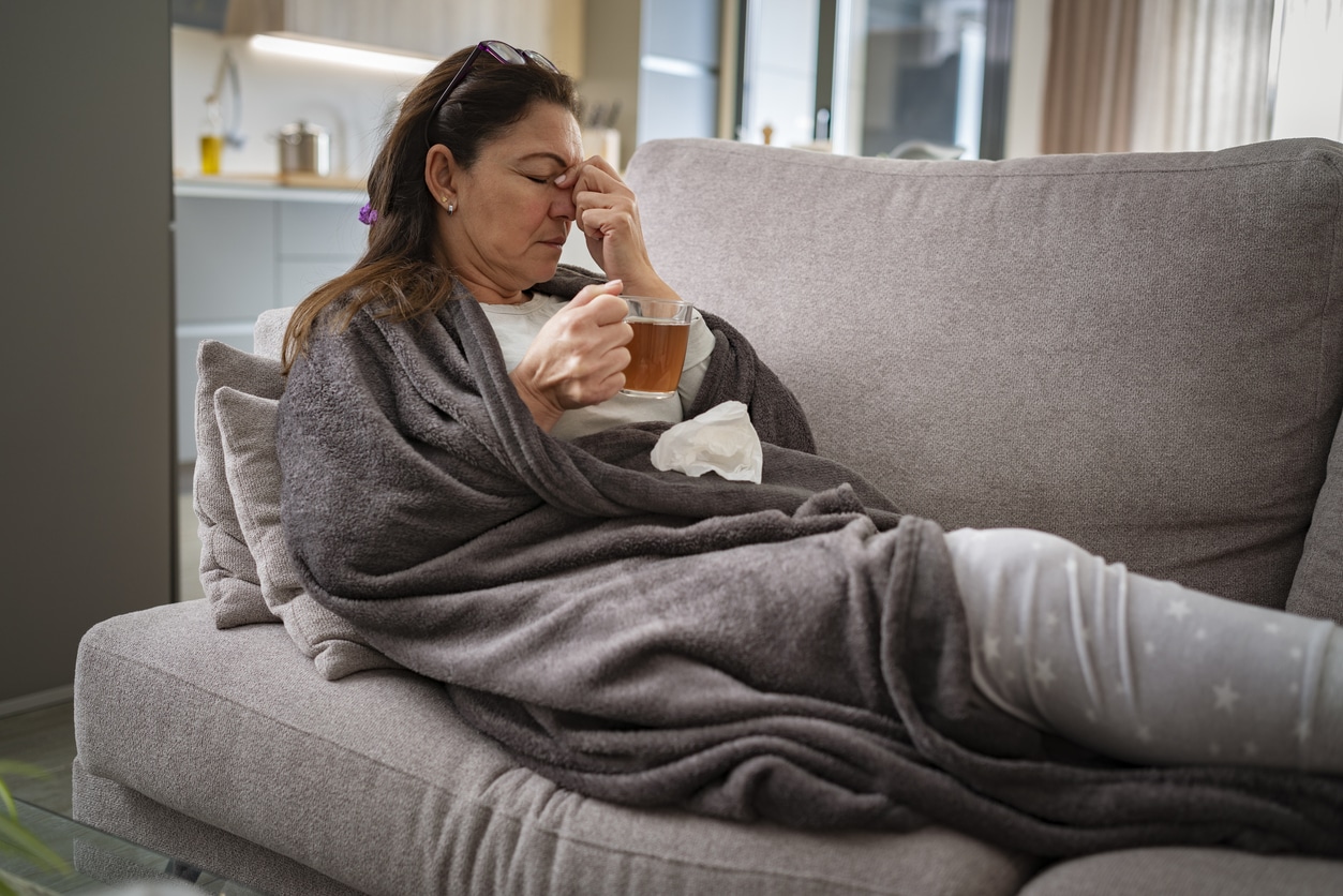 Sick woman resting on sofa drinking hot tea and massaging her nose